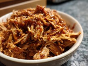 A close-up of shredded slow cooker bbq chicken glistening with sauce in a white bowl.