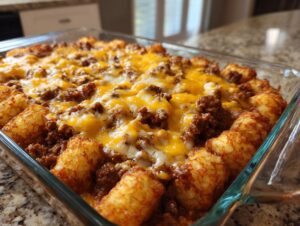 A close-up of a freshly baked Tater Tot Casserole in a glass dish, topped with melted cheddar cheese and seasoned ground beef.