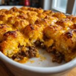 A close-up of a golden-brown Tater Tot Casserole in a white baking dish, with melted cheese oozing over the crispy tater tots and savory ground beef filling.