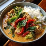 A close-up of a bowl of Thai red curry chicken with rice, featuring tender chicken, broccoli, red peppers, and fresh basil.