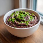 A white bowl filled with thick, dark purple black bean dip, garnished generously with chopped green herbs.