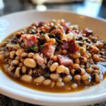 Close-up of a bowl filled with a rich black eyed peas recipe, topped with chunks of smoked ham and herbs.