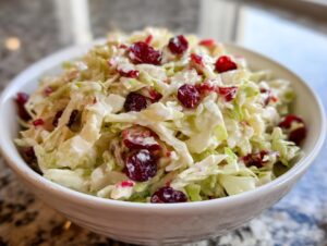 Close-up of a white bowl filled with creamy apple cranberry coleslaw featuring shredded cabbage and dried cranberries.
