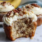 A close-up of a delicious banana cinnamon pecan cupcake with cream cheese frosting and chopped pecans, a bite taken out.