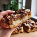A hand holds a delicious slice of Caramel Pecan Turtle Bars, showing layers of coconut, caramel, pecans, and chocolate drizzle.