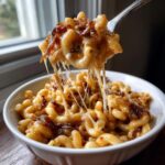 A fork lifts a cheesy, stringy bite of french onion pasta topped with caramelized onions from a white bowl.