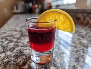 A layered cherry shot cocktail with a bright red bottom and deep purple top, garnished with a slice of orange.