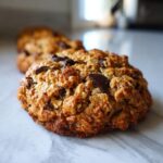 A close-up, macro shot of a thick, chewy chocolate chip oatmeal cookie resting on a white marble surface.