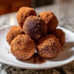 A pile of rich chocolate ricotta truffles generously dusted with cocoa powder on a white plate.