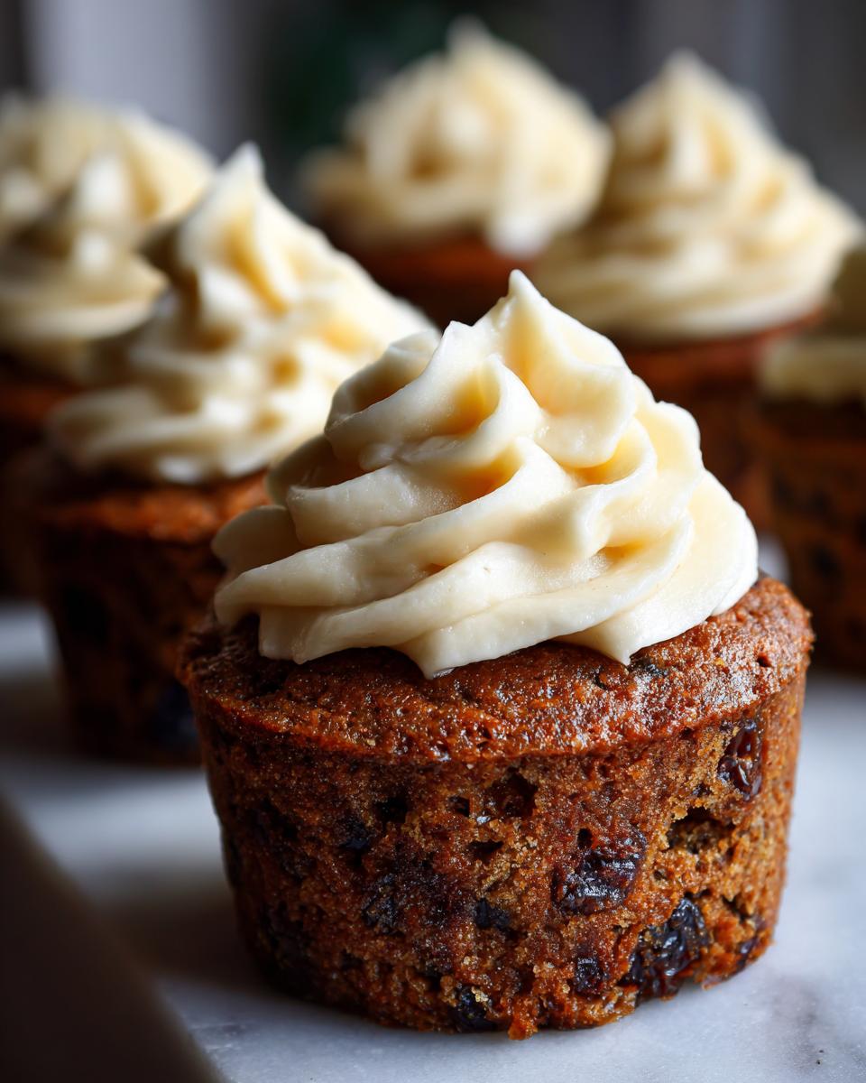 Close-up of a Christmas pudding cupcake topped with swirls of creamy white frosting. The cupcake is rich brown and studded with dried fruit.