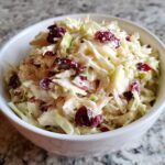 A close-up shot of creamy apple cranberry coleslaw featuring shredded cabbage, apple chunks, and dried cranberries in a white bowl.
