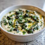 A white bowl filled with creamy dirty martini dip, garnished generously with chopped parsley, black olives, and olive oil.