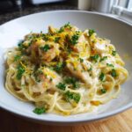 A close-up of a white bowl filled with creamy lemon chicken pasta, garnished with fresh parsley and lemon zest.