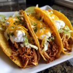 Three crispy chicken tacos filled with shredded chicken, lettuce, cheese, and sour cream, garnished with cilantro and lime.