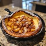 A golden brown Dutch baby pancake with apples, dusted with powdered sugar, served in a cast iron skillet.