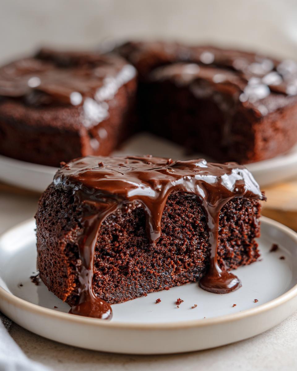 A slice of moist easy chocolate cake generously topped with rich chocolate glaze, with the rest of the cake in the background.