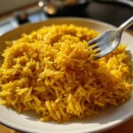 Close-up of bright yellow turmeric rice served on a white plate, with a fork lifting a bite.