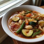 Close-up of a white bowl filled with hearty minestrone soup, featuring zucchini, green beans, carrots, and white beans in a rich tomato broth.