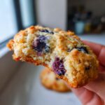 Close-up of a hand holding a bite taken out of a lemon blueberry cookie, showing plump blueberries.