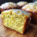 Close-up of a cut lemon poppy seed muffin showing texture and poppy seeds, drizzled with glaze.
