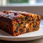 A close-up, tempting slice of moist fruit cake packed with raisins, candied fruit, and nuts, resting on a white plate.