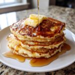 A tall stack of golden brown old fashioned pancakes topped with melting butter and drizzled with syrup.