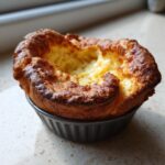 A single, perfectly risen yorkshire pudding with a golden-brown, crispy edge, sitting in a dark muffin tin cup.