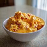 A close-up shot of a white bowl filled with creamy, orange pimento cheese, studded with red pimentos.