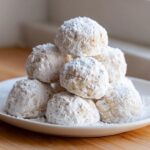 A close-up stack of freshly baked russian tea cakes generously coated in white powdered sugar on a white plate.