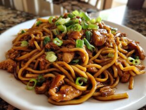Close-up of a plate piled high with glossy Sticky Garlic Chicken Noodles, garnished with fresh green onions.