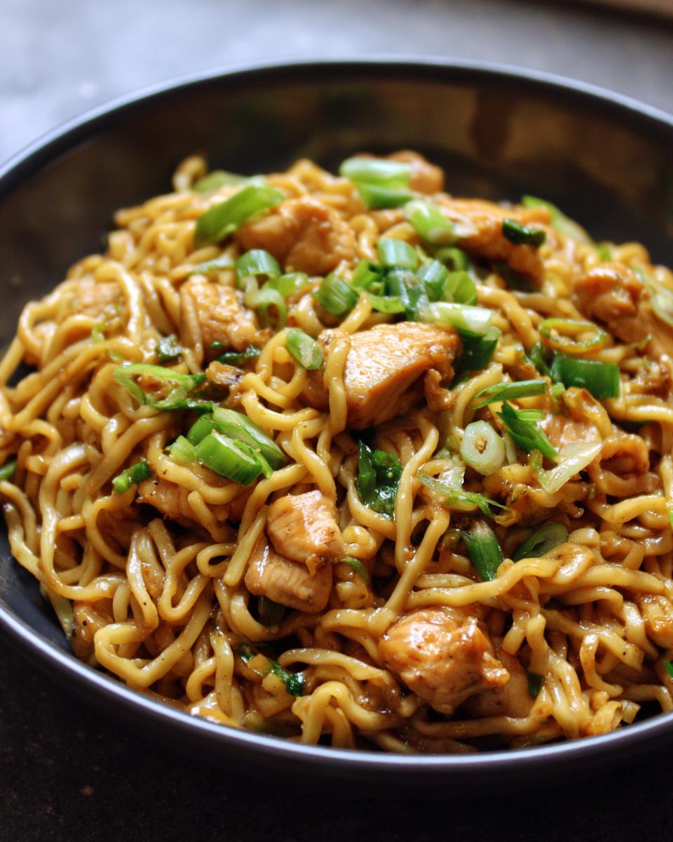 A close-up of a bowl filled with delicious sticky garlic chicken noodles, garnished with chopped green onions.