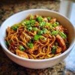 A white bowl filled with delicious sticky garlic chicken noodles, garnished with fresh green onions.