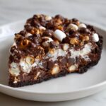A rich slice of texas trash pie loaded with chocolate chips, marshmallows, and pretzels on a white plate.