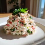 A mound of creamy crab salad mixed with celery and herbs, garnished with fresh parsley on a white plate.