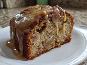 Close-up of a moist slice of apple cake topped with a thick, dripping caramel sauce on a white plate.