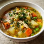 Close-up of a bowl of amazing chicken vegetable soup loaded with shredded chicken, carrots, potatoes, peas, and corn.