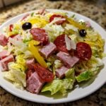 A vibrant plate of grinder salad featuring chopped lettuce, cubed ham, pepperoni slices, yellow peppers, and black olives.