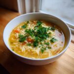 A close-up of a white bowl filled with hot pastina soup, featuring small noodles, carrots, and topped with fresh parsley.