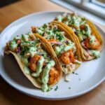 Three fully loaded shrimp taco shells filled with seasoned shrimp, slaw, and drizzled with green avocado crema.
