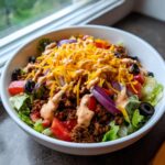 A close-up of a hearty taco salad in a white bowl, featuring seasoned ground beef, shredded cheese, tomatoes, olives, and creamy dressing.