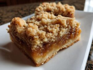 A close-up view of a thick, square apple pie bars slice showing a buttery shortbread base, gooey apple filling, and crumbly topping.