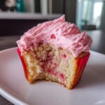 Close-up of a strawberry cupcake with pink frosting, showing a bite taken out to reveal the moist cake and strawberry pieces.