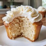 A close-up of a moist vanilla cupcake with a bite taken out, topped with creamy white vanilla frosting.