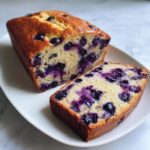 A golden-brown loaf of blueberry banana bread, partially sliced to show the moist interior packed with blueberries.