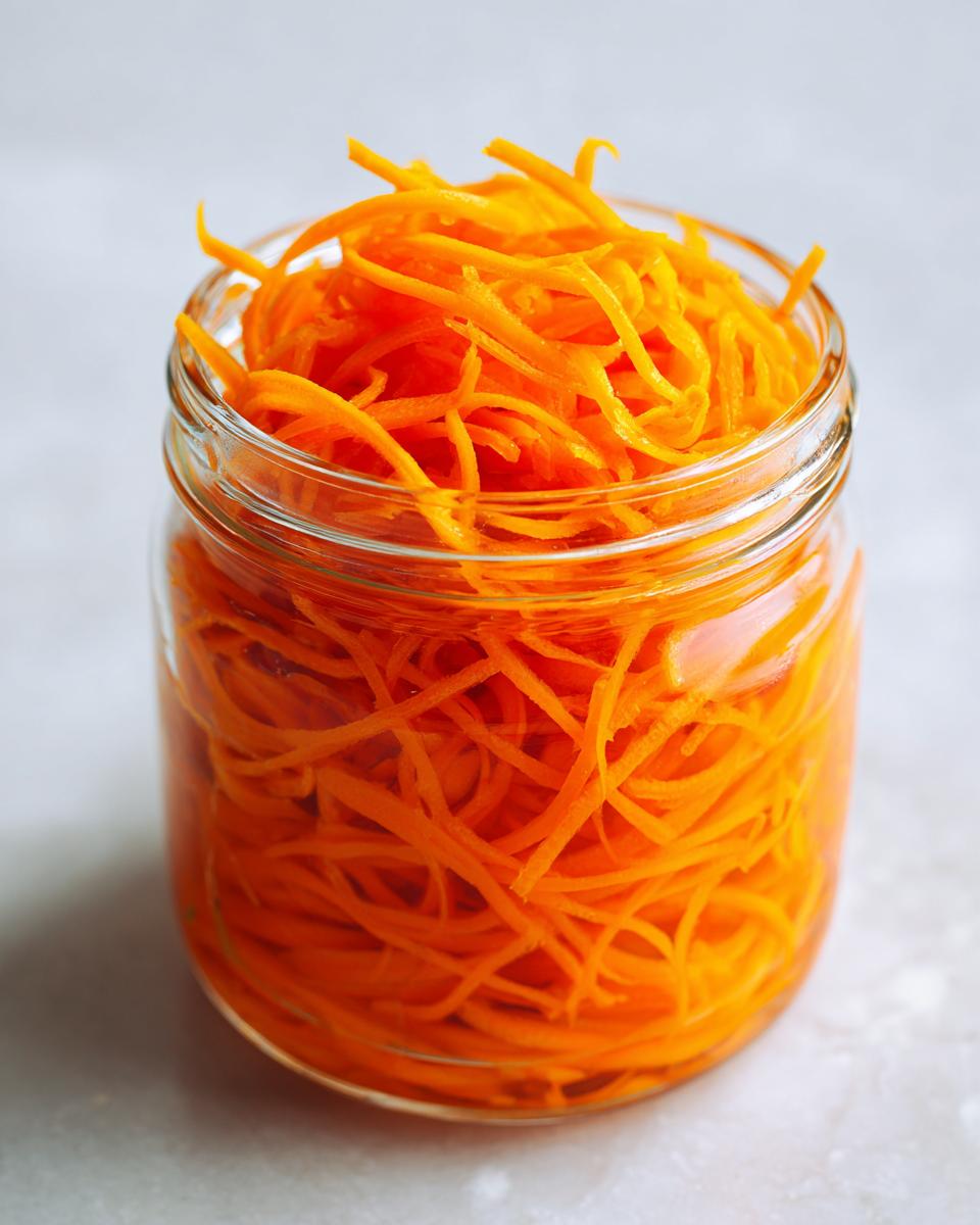 Close-up of bright orange, julienned pickled carrots packed tightly in a clear glass jar.
