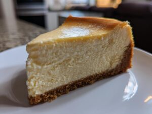 A close-up of a creamy slice of baked cheesecake recipe on a white plate, showing a golden top and graham cracker crust.