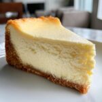 A close-up shot of a thick, creamy slice of cheesecake recipe served on a white plate.