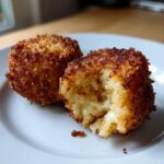 Two golden brown, deep-fried rice balls on a white plate, one cut open revealing a cheesy interior.