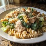 A close-up of a white bowl filled with creamy chicken caesar pasta salad, featuring rotini pasta, chicken chunks, romaine, and croutons.