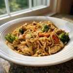 A close-up of a serving of Chicken Chow Mein recipe featuring noodles, chicken pieces, broccoli, and carrots in a white bowl.